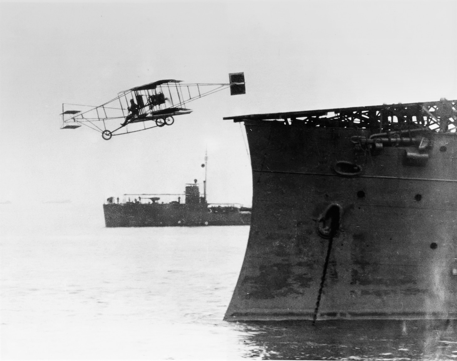 Eugene B. Ely flies his Curtiss pusher airplane from USS Birmingham (Scout Cruiser # 2), in Hampton Roads, Virginia, during the afternoon of 14 November 1910. USS Roe (Destroyer # 24), serving as plane guard, is visible in the background. Photograph from the Eugene B. Ely scrapbook collection, S-005.