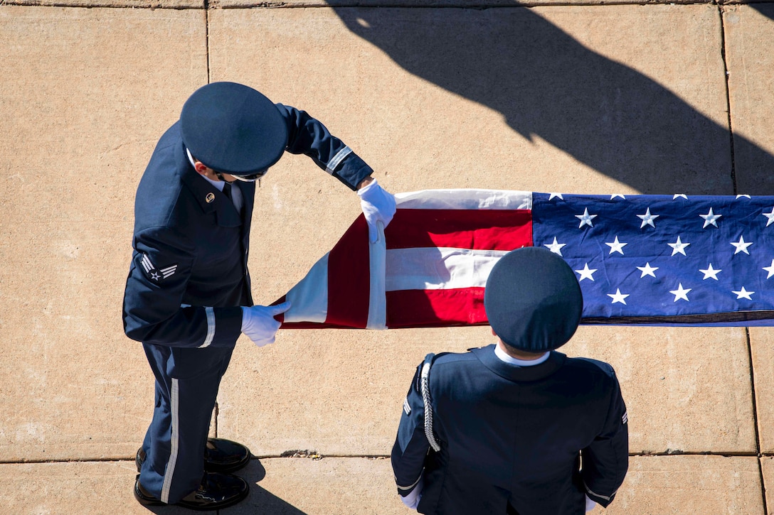 An airman continues folding an American flag as another stands at attention as seen from above.