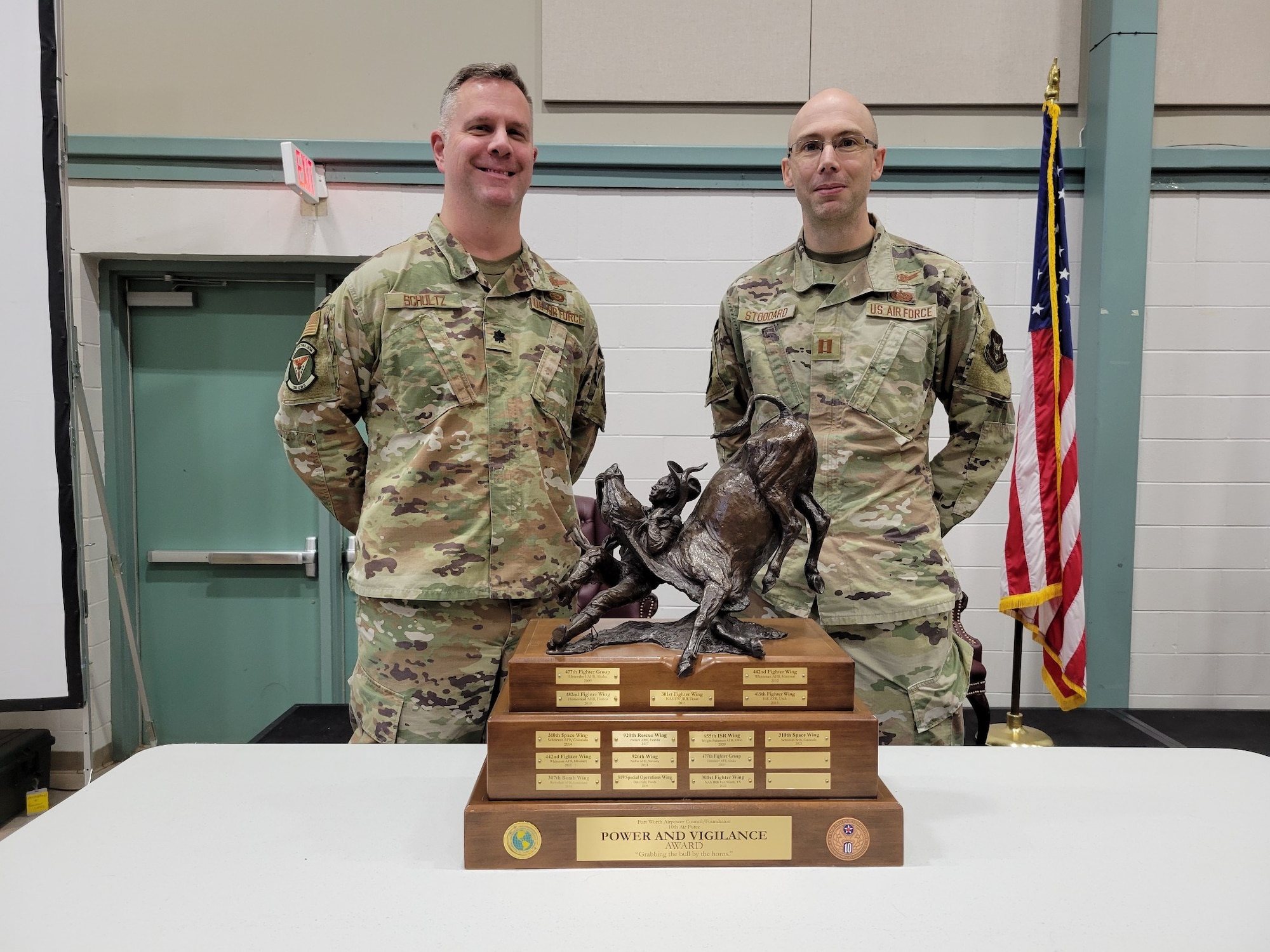 Two people in US Air Force military uniforms stand behind a table with a trophy displayed on top of it.