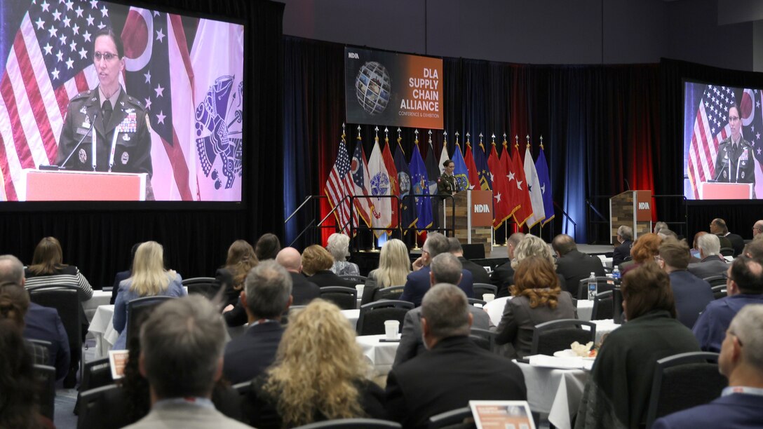 Woman on stage and on screen on left and right speaks to audience.
