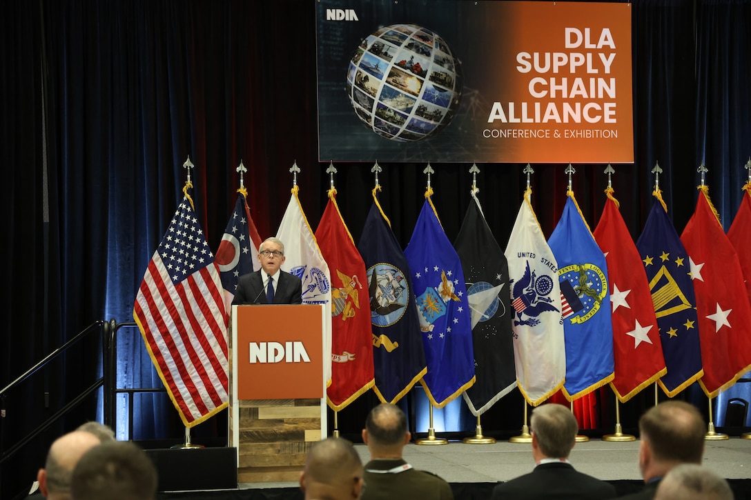 Man stands on stage to the left, flags behind him.