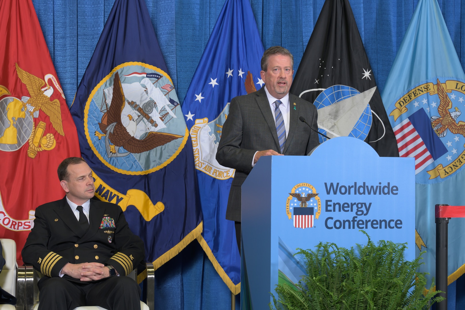 Man in a uniform speaks from a podium as a man in a Navy uniform sits to his side listening.