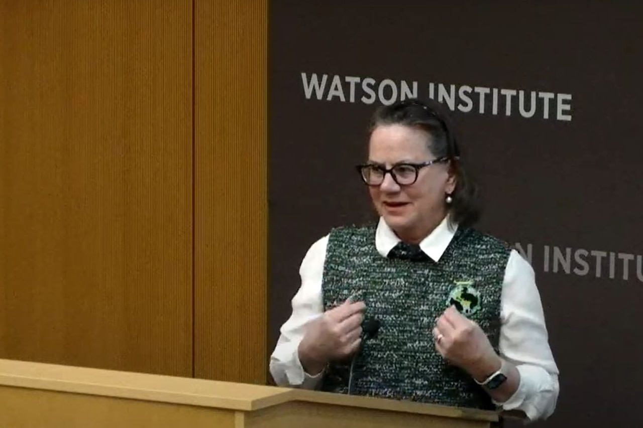 A person in business attire stands at a lectern in front of a sign that reads “Watson Institute.”