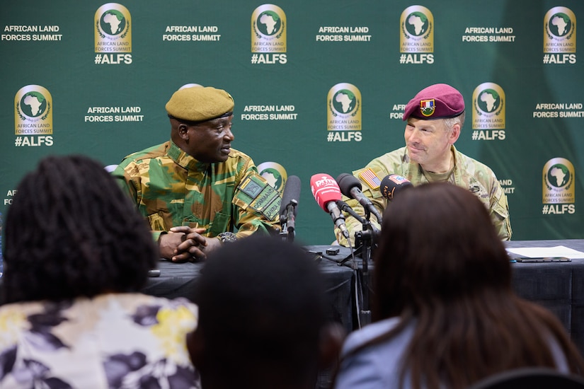 Two men in military uniforms sit next to each other at a table with microphones on the table in front of them. A large banner reading “African Land Forces Summit” is behind them, and three people with their back to the camera are in the foreground.