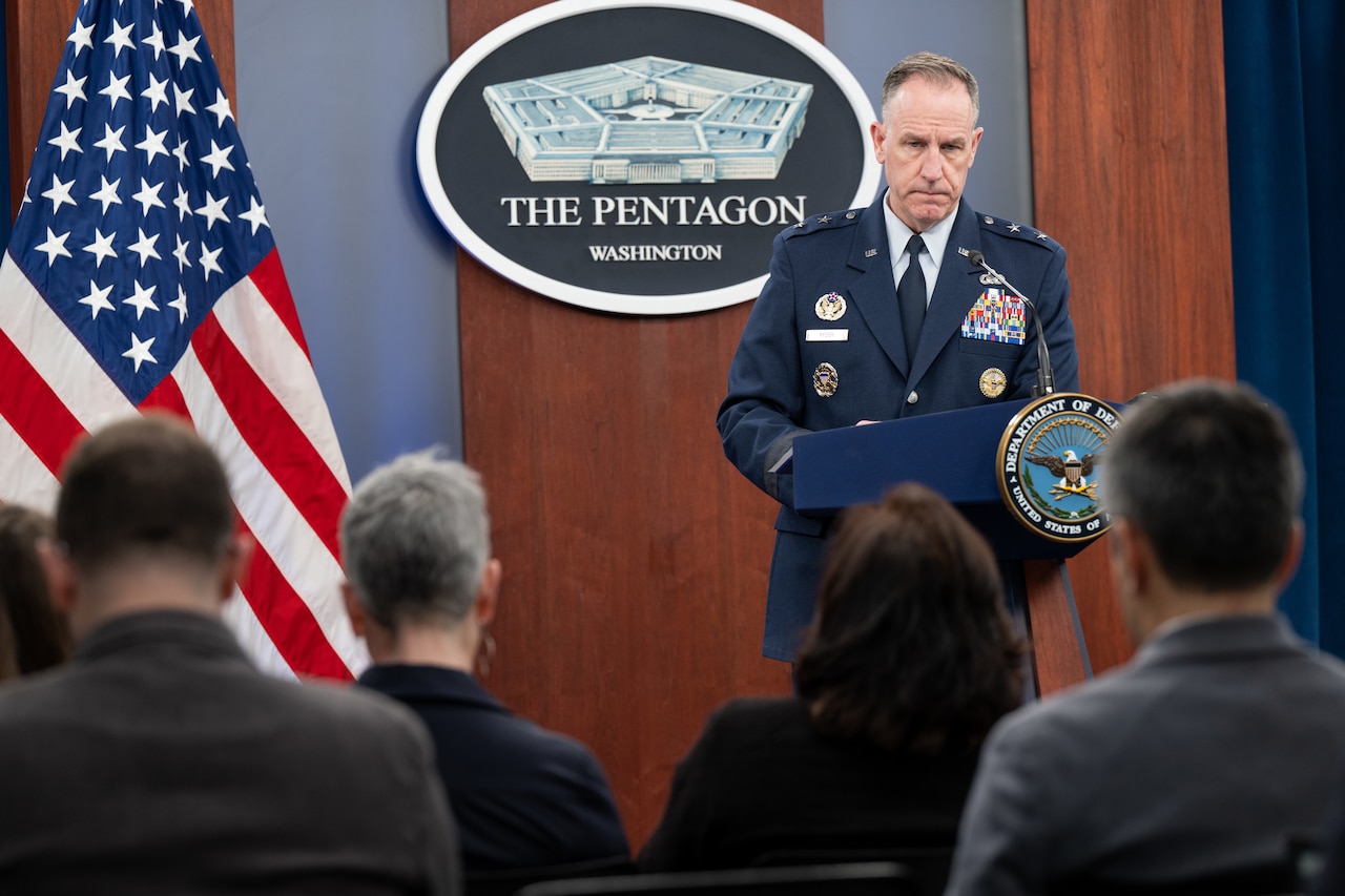 A man in a dress military uniform stands at a lectern addressing people who have their backs to the camera. A Department of Defense logo is behind him on the wall.