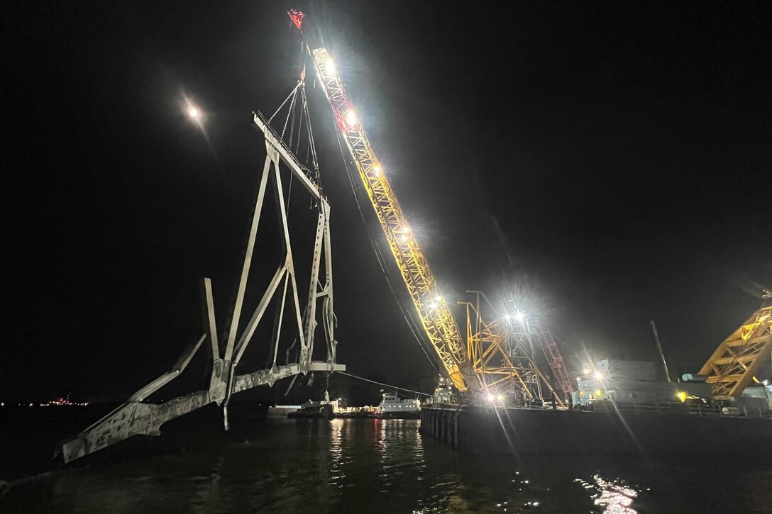 A large crane, lit up at night, lifts a large metal portion of a collapsed bridge from the water.