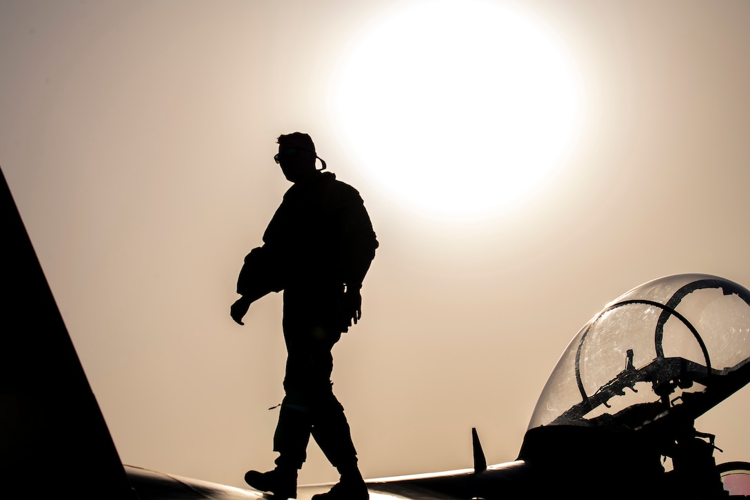 An airman walks across the wing of a military aircraft with a bright sun in the background. The service member is shown in silhouette.