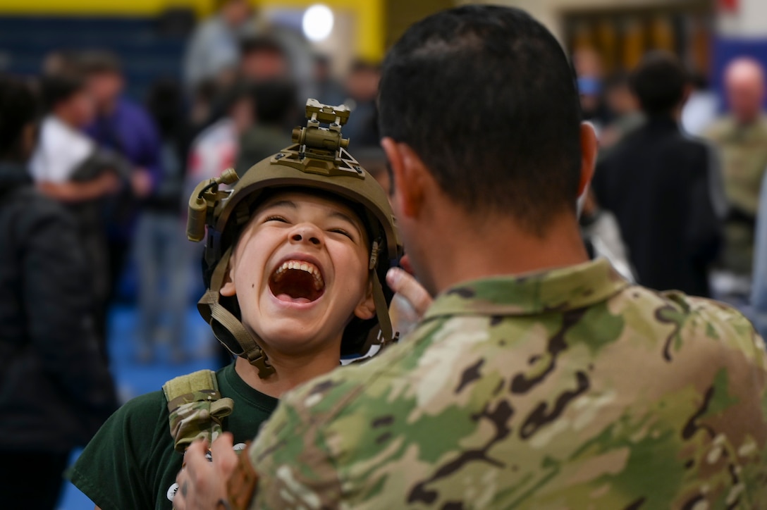 A student laughs while wearing a military helmet as a uniformed service member stands in front of them.