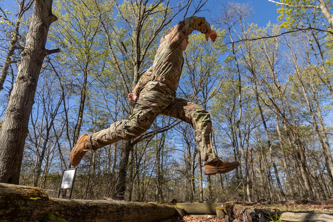 A uniformed service member jumps over an outdoor pit in a wooded area during daylight.