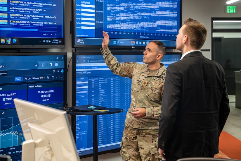 A service member gestures toward a large display screen as another person in business attire watches.