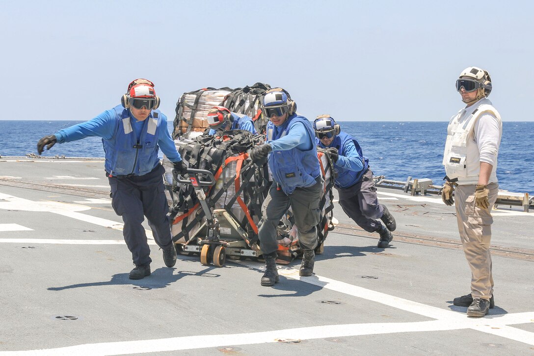 A sailor stands to the right on the flight deck of a ship as four fellow sailors push and pull cargo with a body of water in the background.