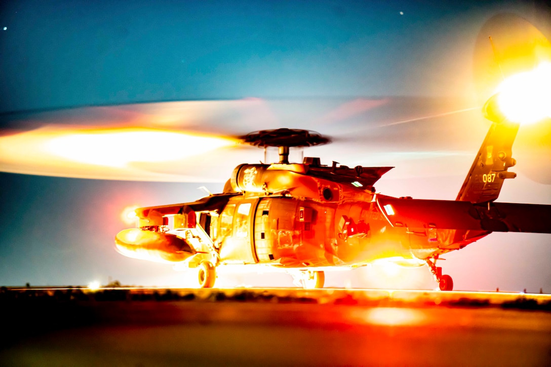 A helicopter’s propeller spins while its parked on a tarmac illuminated by surrounding yellow and red spotlights.