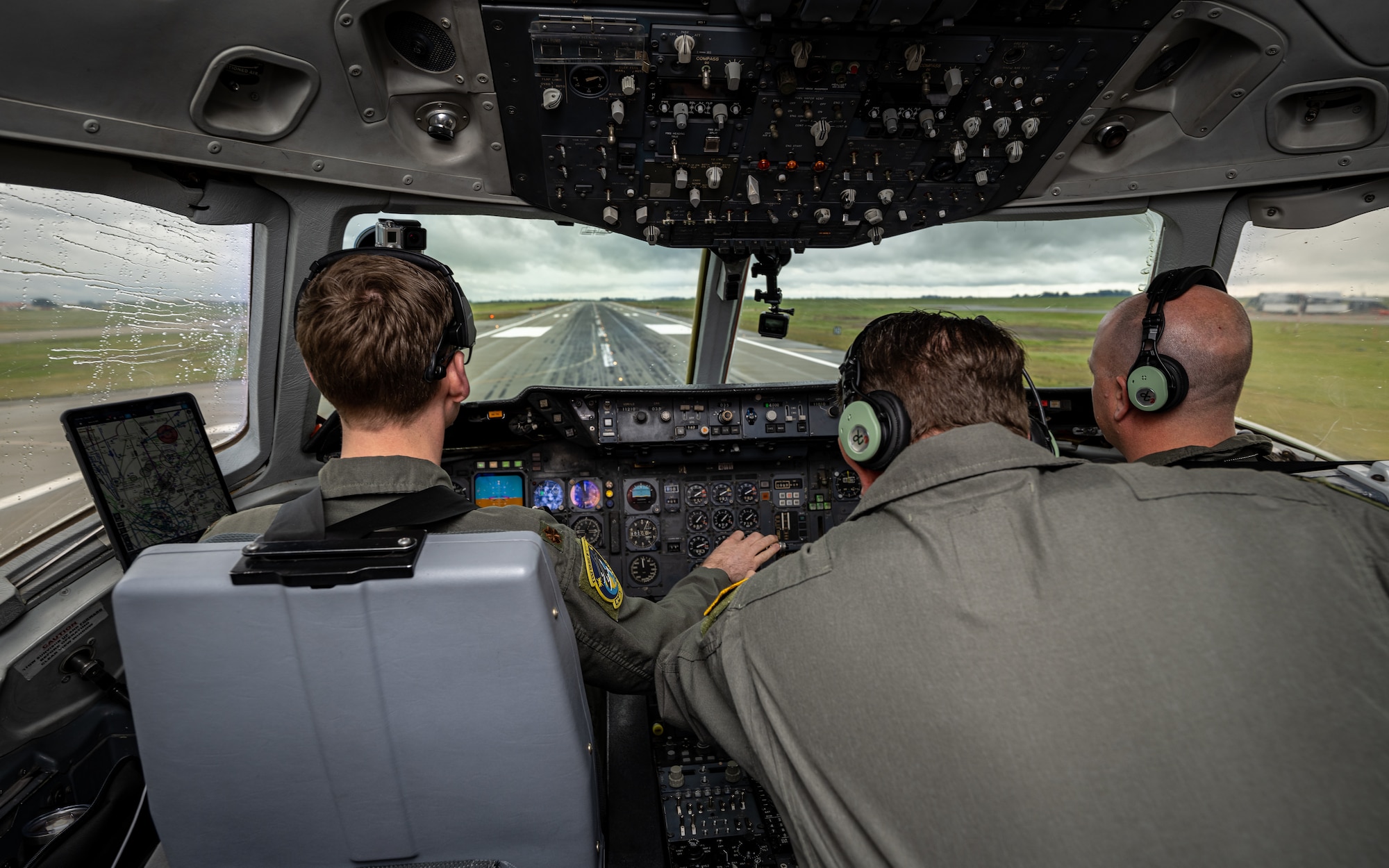Over the shoulder view of 2 pilots and flight engineer looking through cockpit window while taking off with a runway in the background