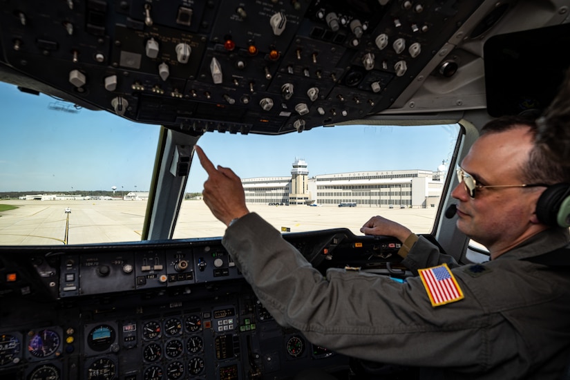 Pilot Lt Col Johnson reaches to press a button inside the cockpit with Wright-Patterson AFB hangars and tower in the background