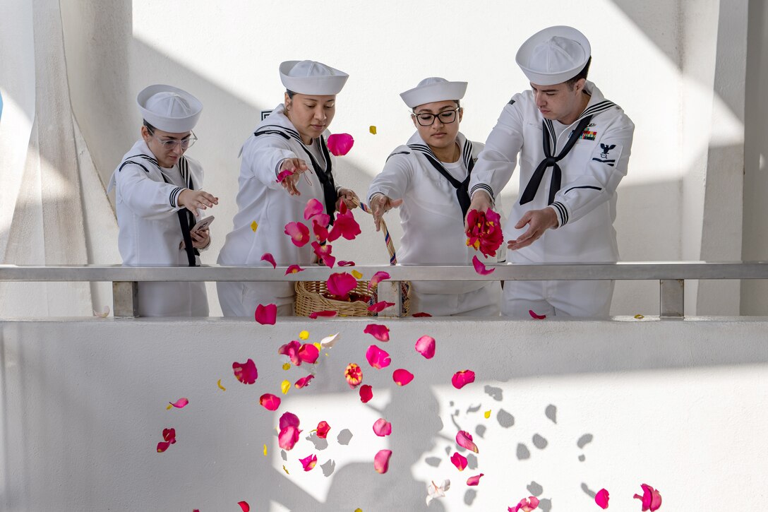 Four sailors drop pink rose petals from a balcony into a well. One sailor holds a wicker basket where the petals are contained.