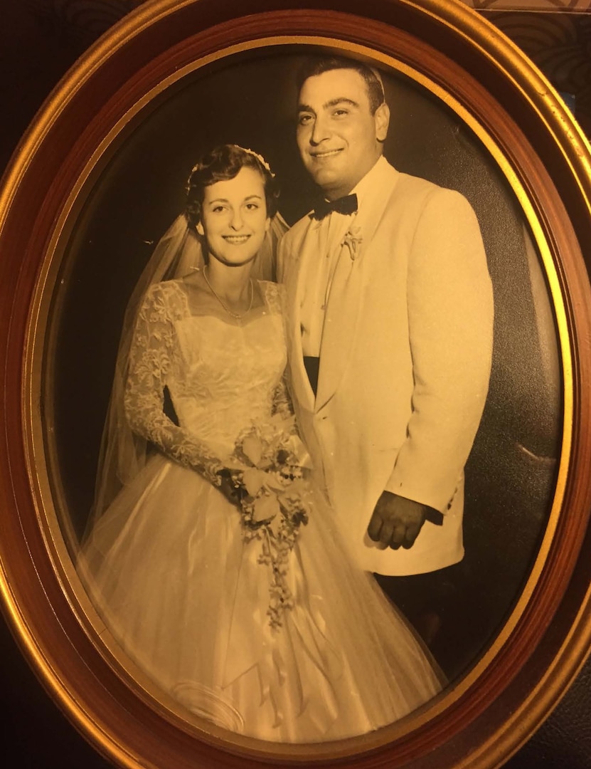 A historical black and white photo of two people on their wedding day. The bride wears a white dress and the groom a white suit. Both are smiling. The bride is holding a bouquet of flowers.