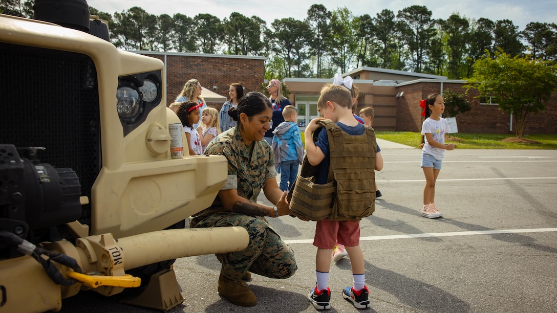 U.S. Marine Corps Cpl. Melina Vazquez, a Woodbridge, Virginia native and mortarman with 2d Battalion, 2d Marine Regiment, 2d Marine Division (MARDIV), assists a child in properly putting on a flak jacket during the annual Month of the Military Child Celebration at Swansboro Elementary School in Swansboro, North Carolina, April 17, 2024. 2d Marine Regiment hosted the display for the annual Month of the Military Child Celebration to foster community relations, while being role models for Onslow County Schools students. (U.S. Marine Corps photo by Pfc. Micah Thompson)