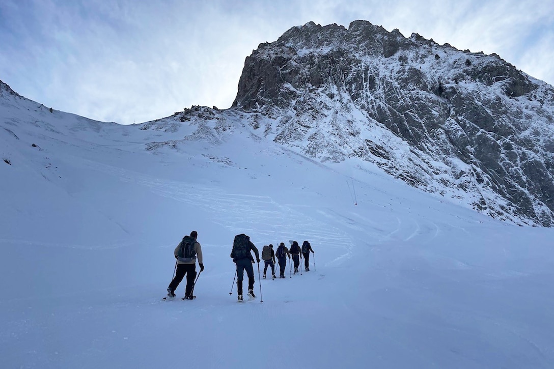 A group of service members use ski poles to hike in snow. A mountain is visible in the background.