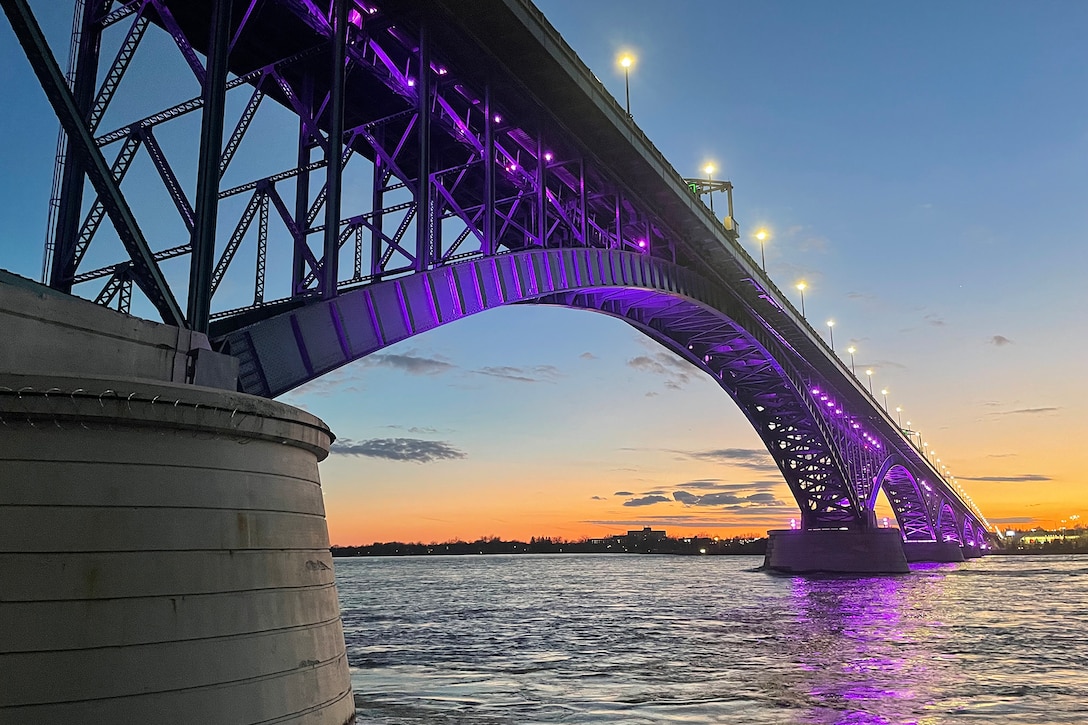 The underside of a bridge over water is illuminated by purple lights at dusk.