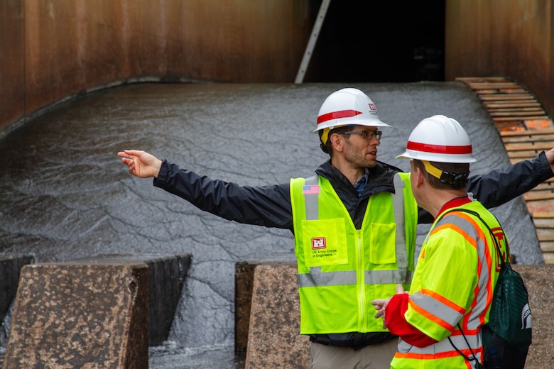 Two men in safety vests and hardhats stand in shallow water and talk with one man pointing both arms.