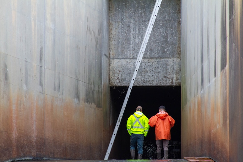 Two men stand in shallow water walk into the opening of a dam with concrete walls on either side.