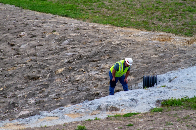 A man in a safety vest and hardhat bends at the waist to inspect a black pipe with rock in the background.
