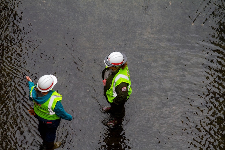 Two women in safety vests and hardhats stand in water and talk.