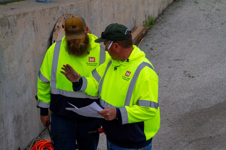 Two men in safety jackets talk and one holds a piece of paper.