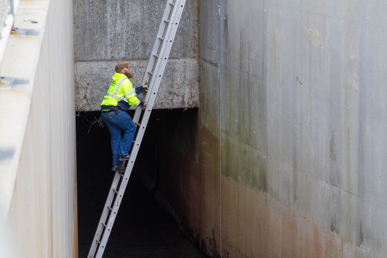 A man in a safety jacket climbs a metal ladder with concrete walls on either side.