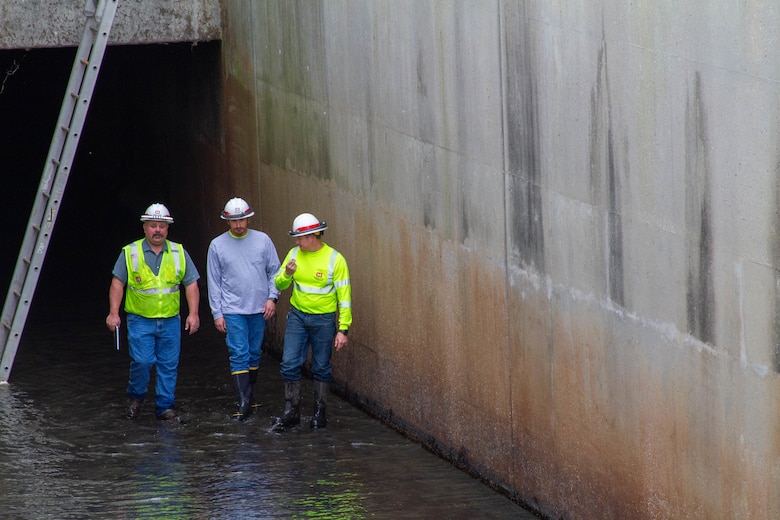 Three men in hardhats stand in water with a metal ladder in the background and brown concrete walls on either side.