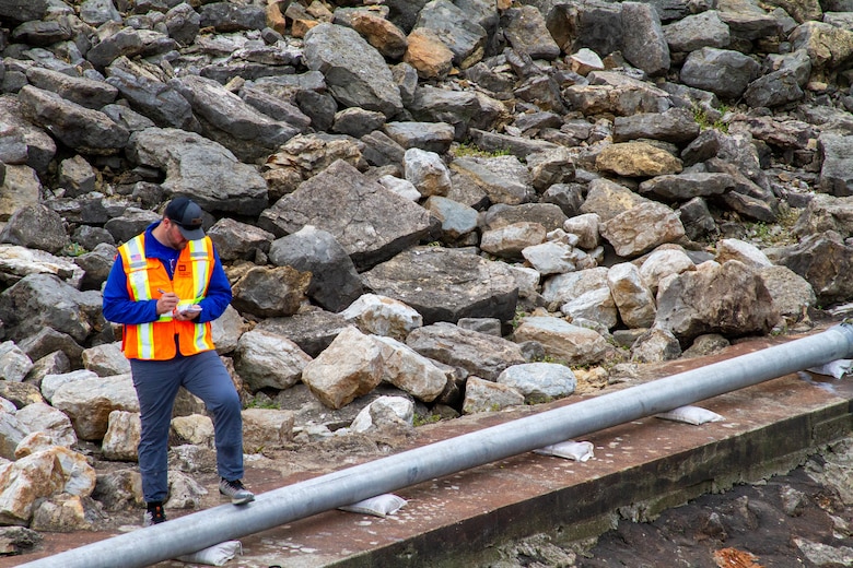 A man in a safety vest stands in a pile of rock and writes on a notepad.