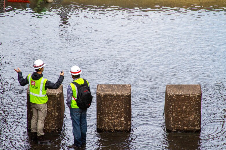 Two men in safety vests stand in water with three concrete blocks in front of them.