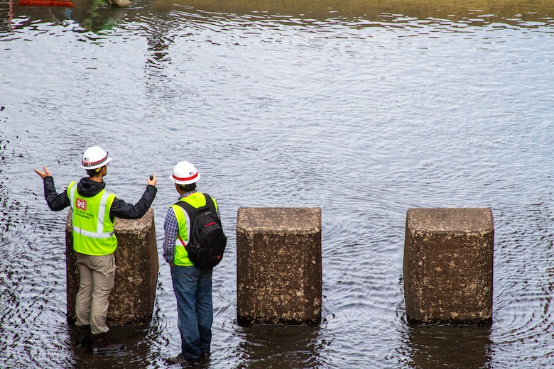 Two men in safety vests stand in water with three concrete blocks in front of them.