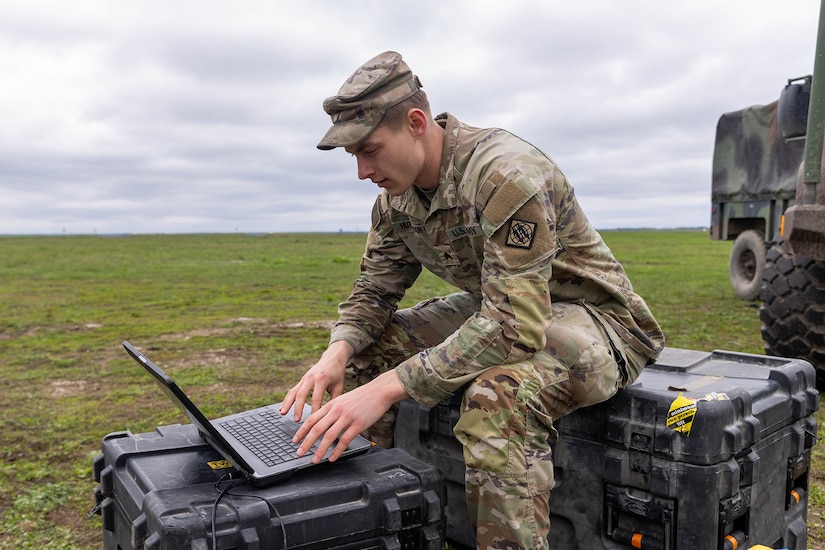 A uniformed service member sits on storage cases outside to type on a laptop.