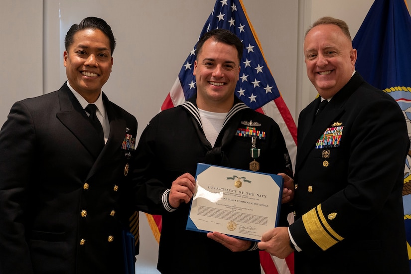 Three service members pose for a photo in front of a U.S. flag. The person in the middle is holding a certificate.
