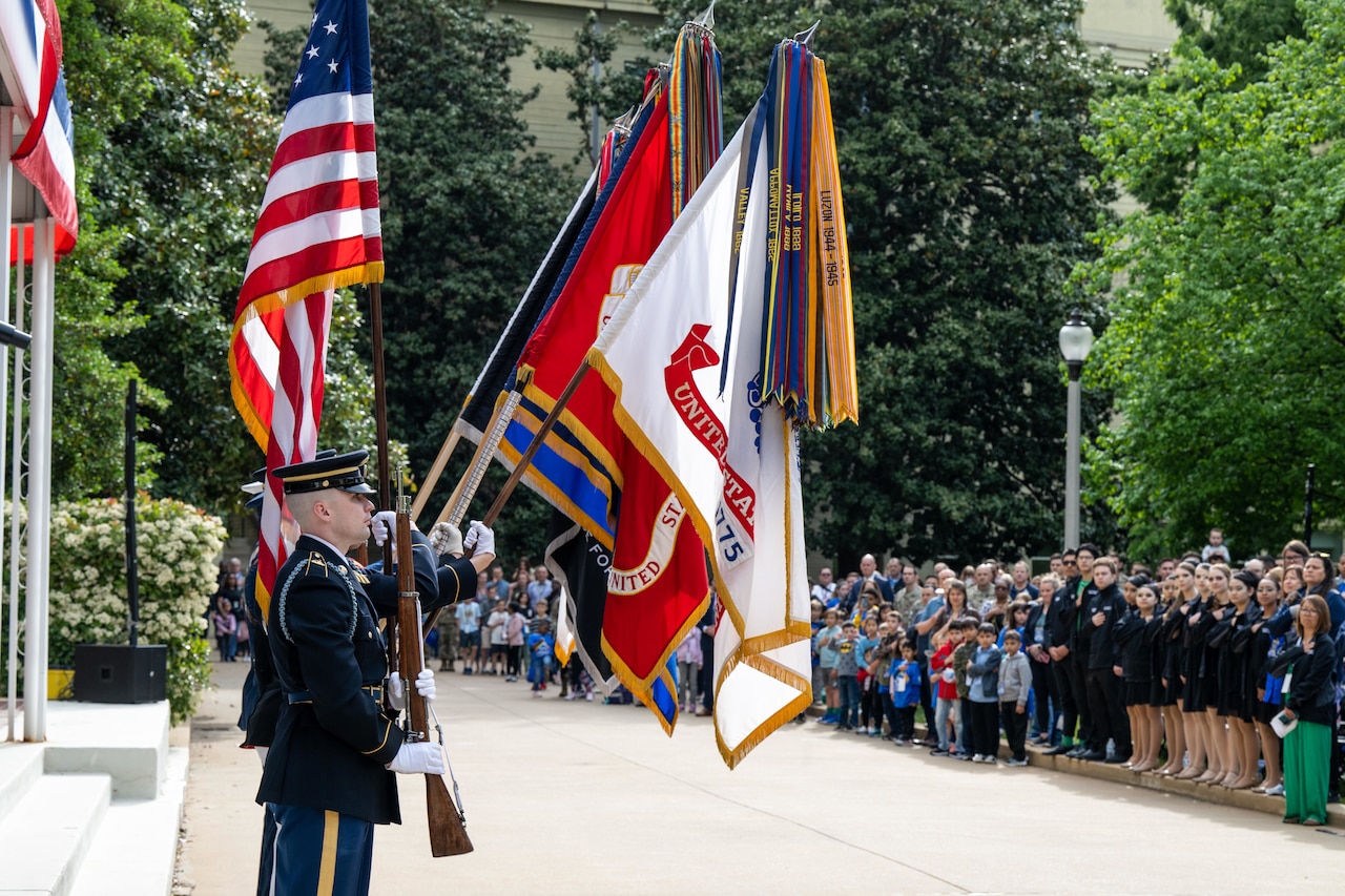Service members holding flags and a ceremonial rifle stand before a large group of people.