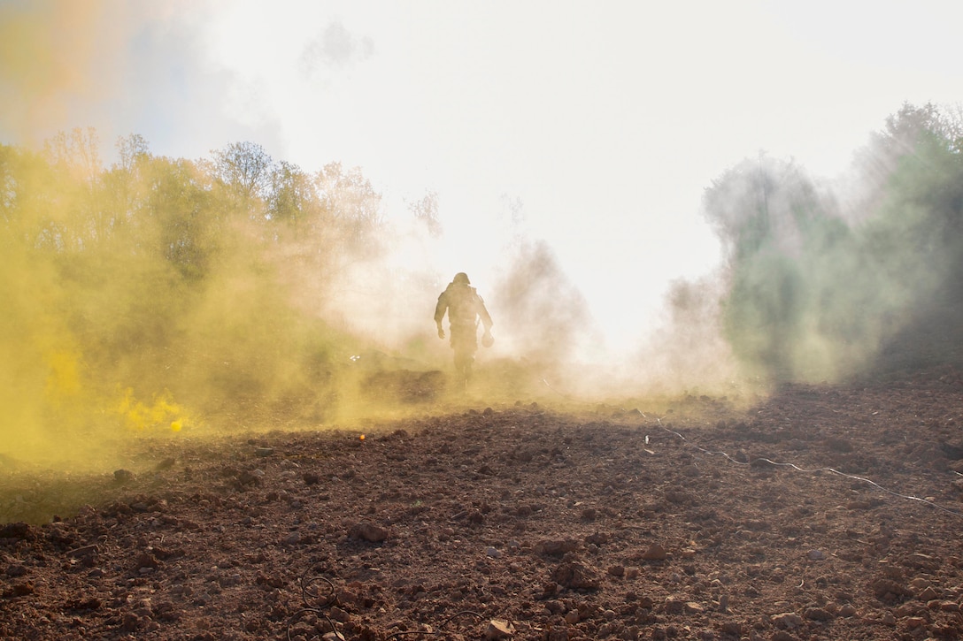 A soldier walks in the dirt through clouds of smoke covering trees in the background.