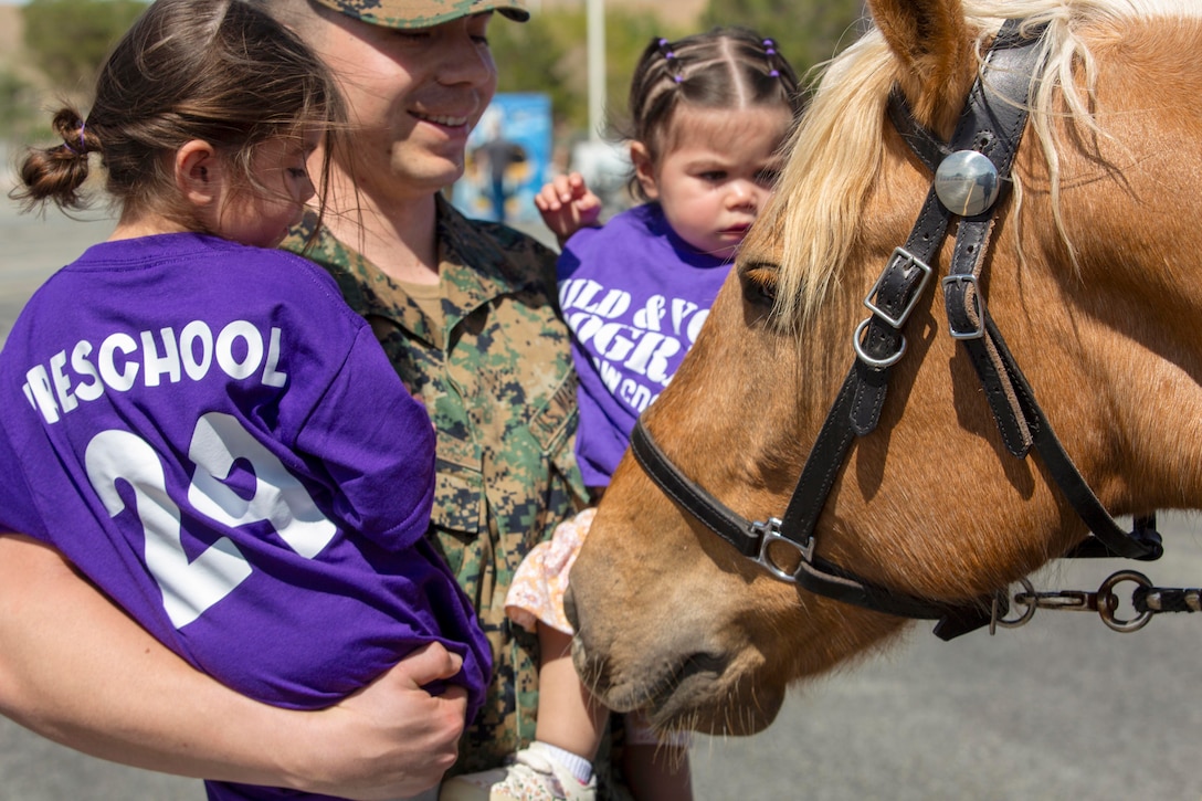 A close-up of a Marine carrying two children while petting a horse.