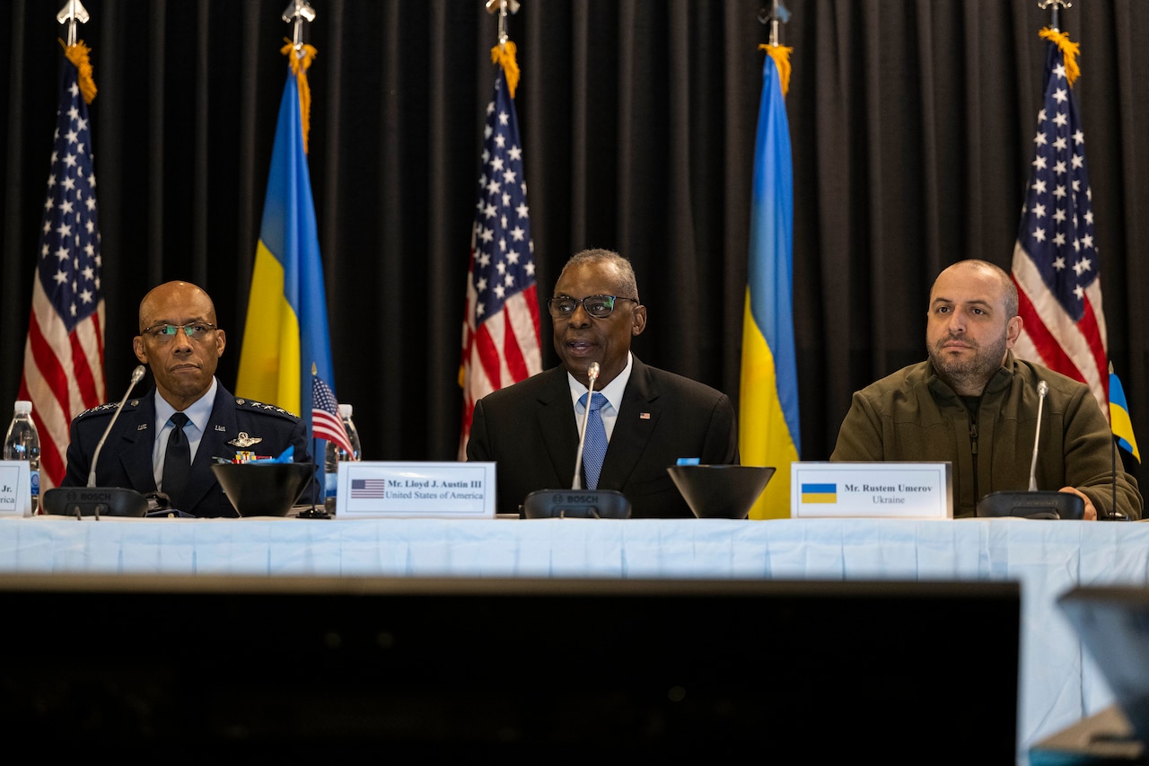 People wearing civilian attire and foreign military uniforms are seated at a table with U.S. and Ukrainian flags in the background.