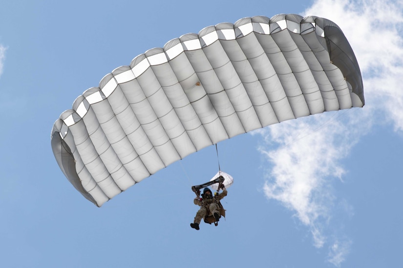 A soldier parachutes to the ground against a blue sky with slight clouds.