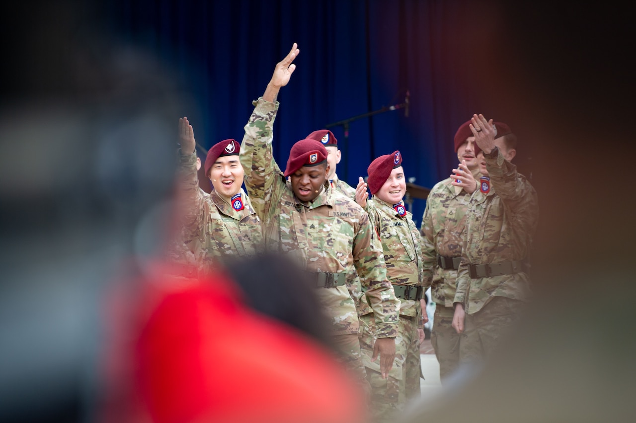 Several soldiers perform, two with raised arms, in front of a very blurred audience and background.