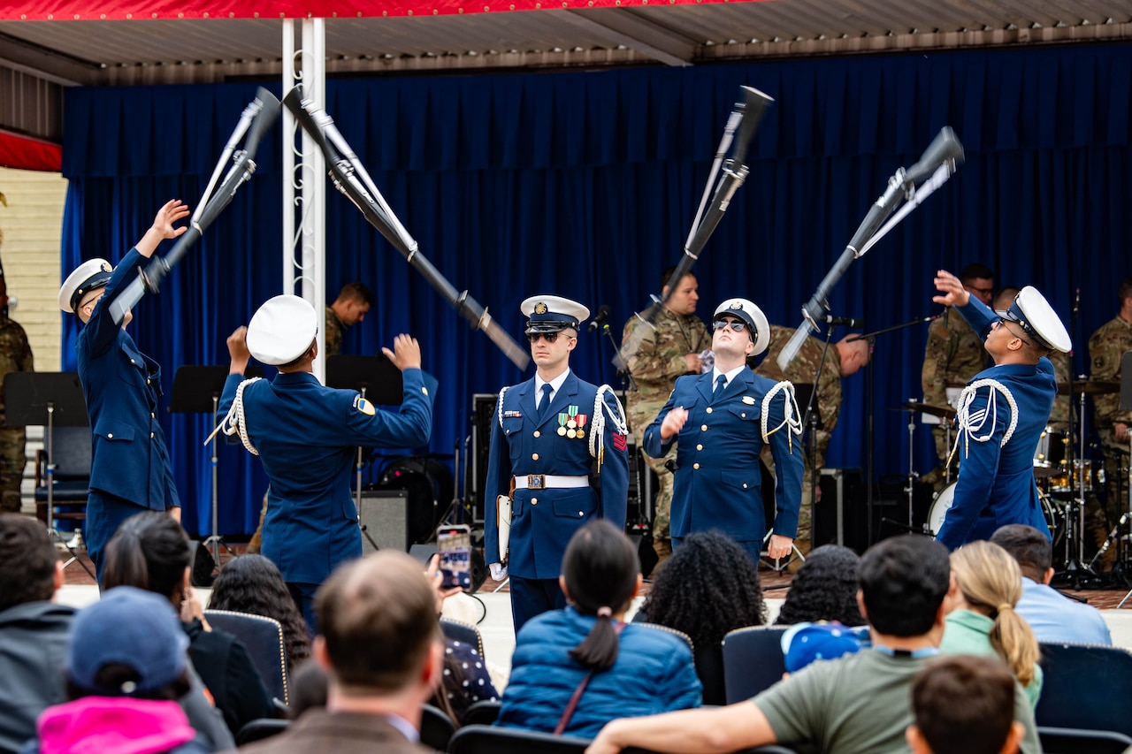Four coast guardsmen toss rifles in the air in front of a stage and a 5th observes while performing for an audience.
