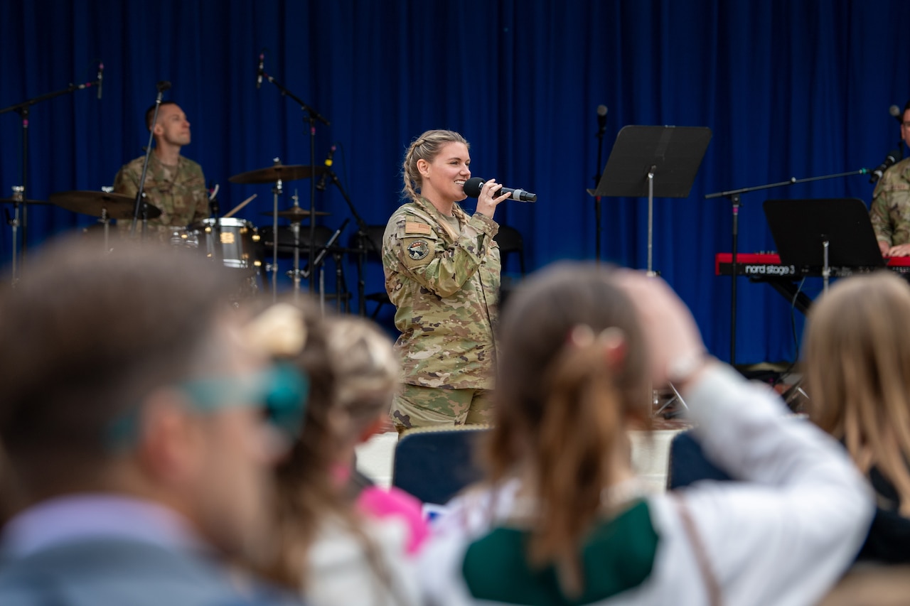 An airman sings on a stage with a drummer behind and a blurred audience in the foreground.