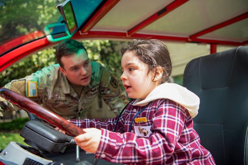 A child sits behind the wheel of a cart and holds on as if to steer while a soldier leans in to give instruction.