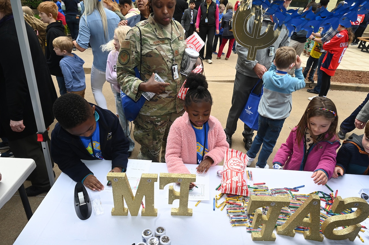 Several children stand at a table with the words SAFFM and other items while a service member stands behind them and others are seen in the background.