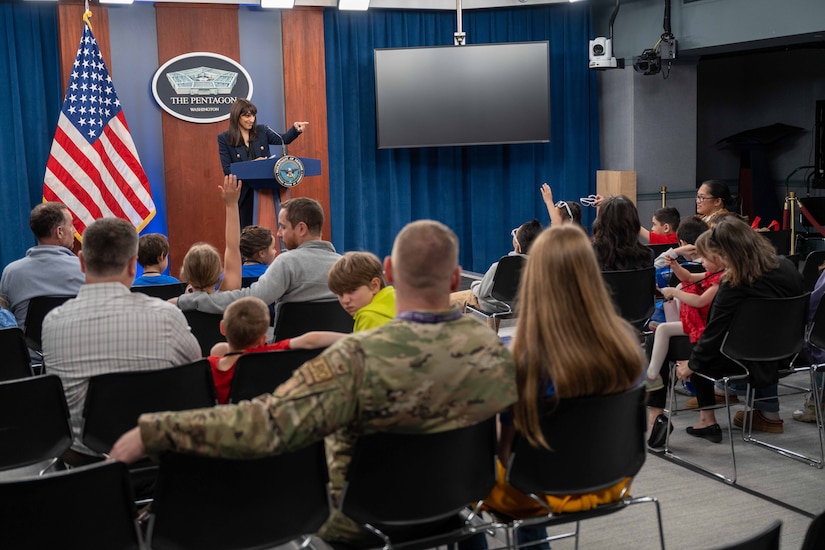 A civilian stands behind a lectern in front of the Pentagon seal and the American flag  and points to children in the audience.