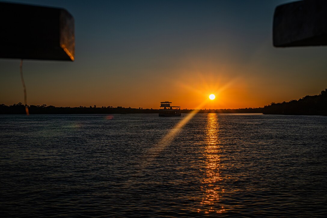 A view of an orange sun setting over open water. A boat and shoreline can be seen in the background.