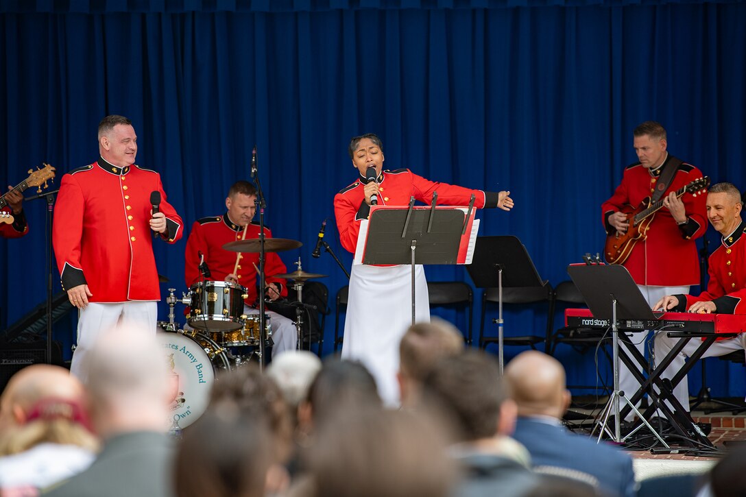 Band members play as a singer gestures. An audience can be seen in the foreground, watching the performance.