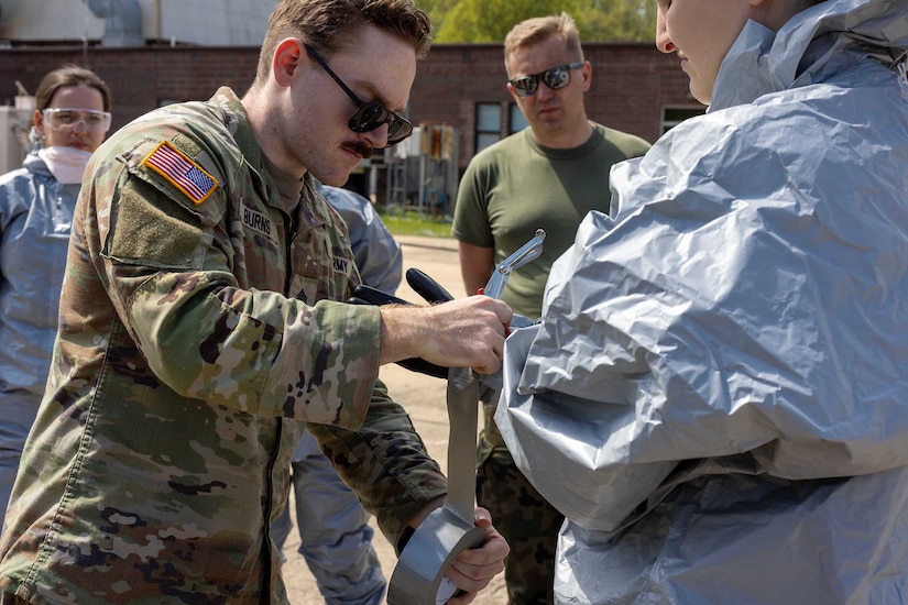 A uniformed service member helps a person in a hazmat suit as others watch.