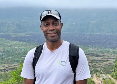 A man stands outside wearing a ball cap, white t-shirt with backpack straps on his shoulders. the photo ends above his waist. behind him is a landscape showing that he is somewhere high above the landscape.
